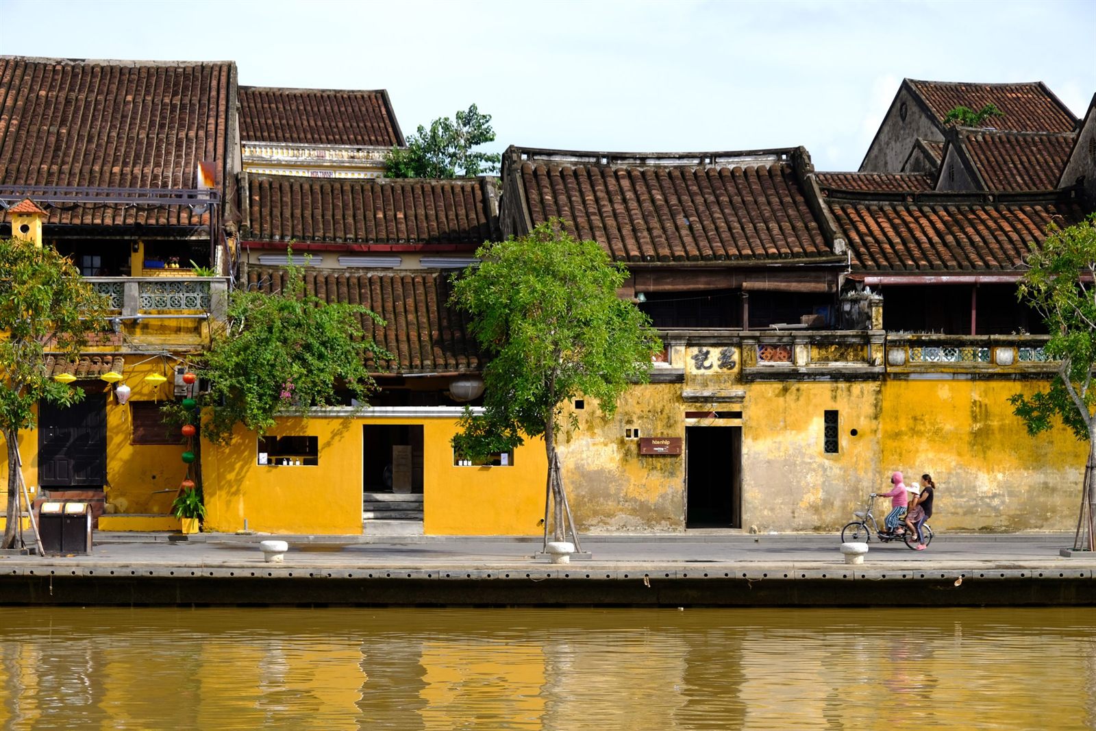Row of old yellow merchant houses with tiled roofs along a quiet riverside street